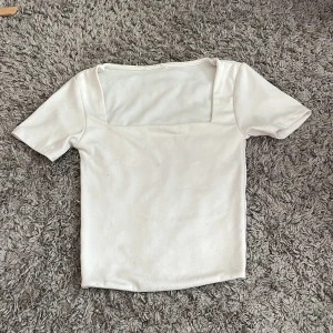 Vit topp med fyrkantig ringning - Säljer en stilren vit topp med korta ärmar och snygg fyrkantig ringning. Toppen är figurnära och gjord i ett mjukt, stretchigt material som känns skönt mot huden. Perfekt att matcha med jeans eller kjol för en clean look.