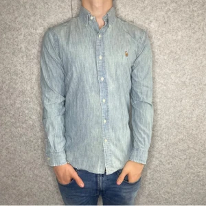 Ljusblå skjorta från Ralph Lauren - Snygg ljusblå skjorta från Ralph Lauren i slim fit-modell. Skjortan är tillverkad i 100% bomull och har klassisk button down-krage, knappar framtill och broderad logga på bröstet. Perfekt för dig som gillar stilrena och tidlösa plagg.