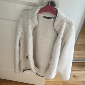 Vit teddyjacka från True North - Mysig vit teddyjacka från True North med långärmad design och fluffigt material. Jackan har en enkel, öppen front och svarta detaljer längs kanterna. Perfekt för att hålla värmen under kyliga dagar.