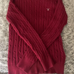 Vinröd kabelstickad tröja från GANT - Vinröd kabelstickad tröja från GANT i stretchig bomull. Klassisk rund halsringning, ribbade muddar och diskret logga broderad på bröstet. Perfekt för dig som gillar en tidlös och stilren look med lite preppy känsla.