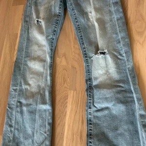 Ljusblå slitna bootcut jeans - Säljer ett par ljusblå jeans med slitningar och hål på benen. Jeansen har bootcut-modell och klassisk femficksdesign. Materialet är jeans i bomull och de har en cool, avslappnad vibe.