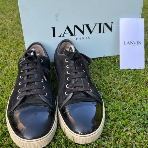 Lanvins - Snygga svarta sneakers från Lanvin med ovandel i mocka och en glansig, lackad tå. Skorna har vita sulor och svarta skosnören med metalländar. Klassisk och stilren design som sticker ut med den blanka tån. Perfekta för dig som gillar exklusiva detaljer. Stygn från sulan har lossnat och även lite gummi som syns på bilderna. Annars bra skick. Pris kan diskuteras