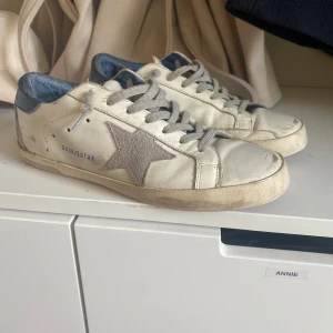 Golden Goose  - Golden Goose sneakers i vitt skinn med grå mockadetaljer och ikonisk stjärna på sidan. Skorna har grå snörning, mörkblå häl och platt sula i gummi. Perfekta för en avslappnad och trendig look. 