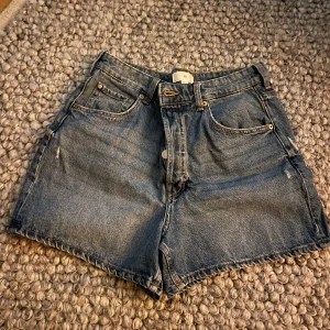 Blå högmidjade jeansshorts H&M - Klassiska blå jeansshorts från H&M med hög midja och raka ben. Shortsen har fem fickor, knappgylf och snygga kontrastsömmar. Perfekta för sommaren och lätta att matcha med allt. Tidlös modell som alltid funkar. En knapp saknades vid köp.
