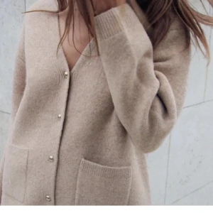 Beige kofta med knappar och fickor - Mysig beige kofta med v-ringning och stora framfickor. Tröjan har långa ärmar, ribbade muddar och stängs med snygga guldfärgade knappar. Perfekt för lager-på-lager och chill dagar.