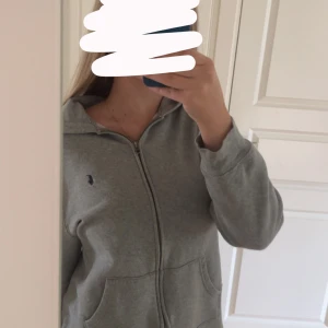 Grå hoodie från Polo Ralph Lauren - Säljer en grå hoodie från Polo Ralph Lauren med dragkedja och huva. Tröjan har långa ärmar, två fickor framtill och den klassiska broderade loggan på bröstet. Perfekt för chill dagar eller när du vill ha en clean och enkel look. Skulle säga att storleken är som S