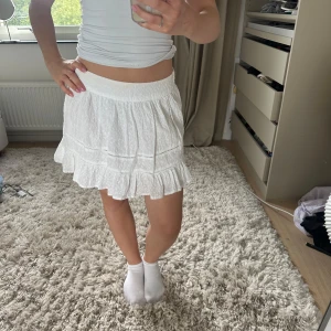 Vit volangkjol med broderade detaljer - Supersöt vit kortkjol med volangkant och broderade detaljer. Kjolen har resår i midjan för en bekväm passform och är gjord i ett lätt bomullstyg. Perfekt för varma dagar och ger en fräsch och somrig vibe.