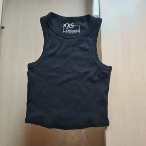 Svart ribbad linnetopp - En svart, ribbad linnetopp från FSBN Sister i storlek XXS. Toppen är ärmlös med breda axelband och rund halsringning. Perfekt för en enkel och clean look, lätt att matcha med allt. Materialet är mjukt och stretchigt för en bekväm passform. Går att köpa 3-pack (kolla mina annonser)