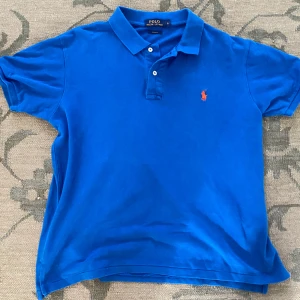 Blå pikétröja från Polo Ralph Lauren - Klassisk blå pikétröja från Polo Ralph Lauren i bomull med orange broderad logga på bröstet. Nypris 900kr