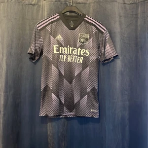 Olympique Lyonnais Adidas matchtröja - Snygg Olympique Lyonnais fotbollströja från Adidas med svart och lila geometriskt mönster, korta ärmar och klassiska Adidas-ränder på axlarna. Tillverkad i återvunnen polyester, lätt och andas. Emirates Fly Better-tryck på bröstet och klubbmärke på vänster sida.