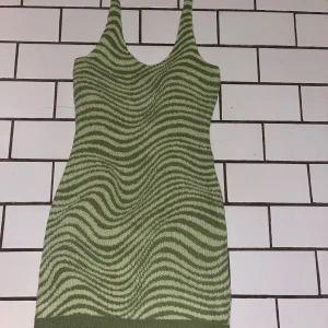 Grön stickad kortklänning med mönster - Säljer en grön och ljusgrön stickad kortklänning med vågigt randigt mönster. Klänningen har smala axelband att knyta i nacken och ärmlös design, perfekt för dig som gillar en unik och trendig look. Materialet är mjukt och stretchigt stickat, vilket gör den bekväm att bära.