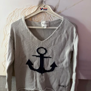  Grå  tröja med ankare Bondelid - Stickad tröja från Bondelid i grått  med stort  blått ankare framtill. V-ringad hals och långa ärmar. Perfekt för en nautisk look och chill vår- eller sommarkväll. Mjuk och skön känsla, klassisk Bondelid-brodyr nertill.Har även en marinblå 