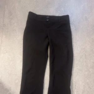 Svarta kostymbyxor med raka ben - Snygga svarta kostymbyxor med raka ben och två knappar i midjan. Byxorna har bälteshällor och bakfickor med lock och knapp. Perfekta för en clean och stilren look. Passar till skjorta eller t-shirt beroende på humör.