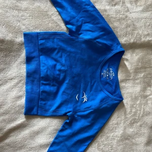 Blå croppad tröja från Peach Tech - Snygg blå croppad tröja från Peach Tech med lång ärm och rund hals. Tröjan har ett stilrent tryck framtill och är gjord i ett stretchigt material av nylon och elastan. Perfekt för träning eller chill dagar. Ribbad kant nedtill ger extra cool vibe.