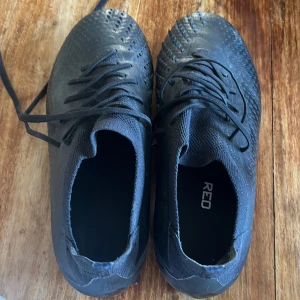 Svarta fotbollsskor från Adidas strl 40 - Säljer ett par svarta fotbollsskor från Adidas i storlek 40. Skorna har snörning, mönstrad ovandel och robusta dobbar för bra grepp på planen. Tillverkade i syntetmaterial med sportig look och diskret Adidas-logga på sulan.