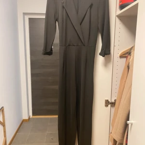 Svart jumpsuit med kavajkrage och bälte - Snygg svart jumpsuit med lång ärm, kavajkrage och knytbälte i midjan. Raka ben och uppvikta ärmslut ger en clean och modern look. Perfekt för dig som gillar stilrena plagg med lite edge. Materialet känns mjukt och följsamt.