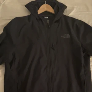 Svart vindjacka från The North Face L - Svart vindjacka från The North Face i storlek L. Jackan har huva, hel dragkedja framtill och diskret logga på bröstet. Perfekt för blåsiga dagar och enkel att matcha med allt. Snygg och funktionell design för dig som gillar streetwear.