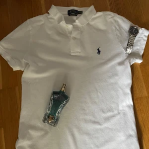 Vit Ralph Lauren piké  - Vit kortärmad polo från Ralph Lauren i modellen The Skinny Polo. Klassisk piké med krage, två knappar och den ikoniska broderade loggan i marinblått på bröstet. Tillverkad i mjuk bomull som andas och har en slim passform. Vid frågor är det självklart bara att höra av dig!😊