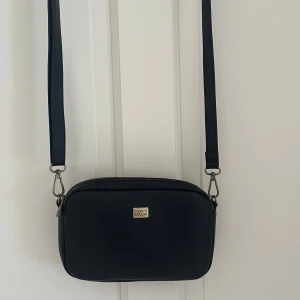 Svart axelväska från Pipol's Bazaar - Stilren svart axelväska från Pipol's Bazaar i skinnimitation med justerbar rem och dragkedja upptill. Väskan har ett huvudfack med innerficka och en mindre ficka på baksidan. Perfekt storlek för mobil, plånbok och nycklar. Pris går att diskuteras