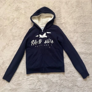 Mörkblå zip up Hollister hoodie med foder - Mysig mörkblå zip up hoodie från Hollister med vit broderad logga och text på bröstet. Tröjan har huva med vitt fluffigt mjukt foder, dragkedja framtill och två fickor. Perfekt för kyliga dagar!🤍