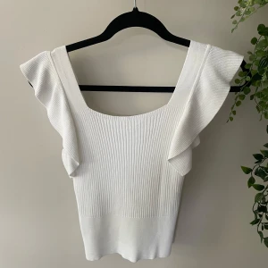 Vit ribbad topp med volangärmar - Supersöt vit ribbad topp med breda axelband och volangdetaljer på axlarna. Toppen har en fyrkantig ringning och är figurnära i modellen.  Den är köpt i Spanien och alldrig använd pga för liten. Storlek vet jag ej men skulle säga xs/s. Pris går att diskutera! 