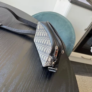 Grå och svart Dior axelväska - Snygg axelväska från Dior med klassiskt monogrammönster i beige och svart. Väskan har två fack med dragkedja, justerbar axelrem och detaljer i svart textil och metall. Perfekt storlek för det viktigaste och en riktigt trendig accessoar. Liten skavank då ena dragkedjan saknar krok. Köpte väskan ny på dior.com för 14000 kr.