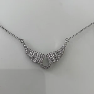 Silverhalsband med glittriga vingar - Snyggt zadig liknande halsband i silverfärg med ett hängsmycke format som två vingar. Vingarna är täckta av små glittrande stenar på framsidan och har en slät baksida. Kedjan är tunn och har ett klassiskt karbinlås. Perfekt för dig som gillar unika detaljer och lite extra bling.