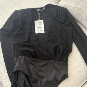 Svart glittrig omlott bodysuit - Svart långärmad bodysuit från TTRI med glittrigt tyg upptill och omlott-skärning. Nedre delen är i slät, stretchig polyester. Perfekt för dig som vill ha en snygg och bekväm topp med lite extra glitter.