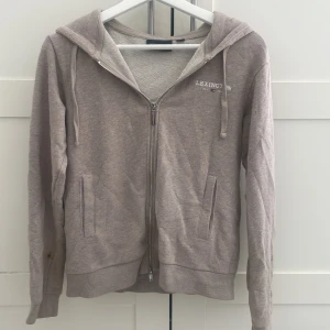 Beige hoodie från Lexington med dragkedja - Säljer en beige hoodie från Lexington med broderad logga på bröstet. Tröjan har huva, snörning och praktisk dragkedja framtill samt två fickor. Materialet är mjuk bomull och modellen är normal passform med långa ärmar. Perfekt för chill dagar och enkel att matcha.