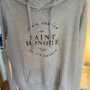 Ljusgrå hoodie från Cotton On - Säljer en ljusgrå hoodie från Cotton On med tryck 'Saint Honoré Paris France' på bröstet. Tröjan har huva med snörning, magficka och långa ärmar. Perfekt för chill dagar och enkel att matcha med jeans eller joggers.