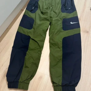 Nike gröna och svarta täckbyxor - Snygga täckbyxor från Nike i grönt och svart med coola paneler och resår vid benslut. Byxorna har dragkedjefickor framtill och en vit Nike-logga på låret. Perfekta för dig som vill sticka ut med sportig stil.