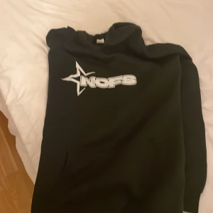 Svart NOS hoodie med tryck, stl M - Svart hoodie från NOS i storlek M med stort vitt tryck och stjärndetalj på bröstet. Klassisk huva och känguruficka framtill. Tillverkad i mjuk polyester, perfekt för chill eller streetwear. Snygg och enkel att matcha med andra plagg.
