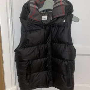 Svart dunväst från Burberry - Säljer en svart dunväst från Burberry med klassiskt rutigt foder i grått, vitt och rött. Västen har dragkedjor på fickorna, knappar framtill och en hög krage. Perfekt för lager-på-lager och riktigt snygg med den ikoniska Burberry-looken.