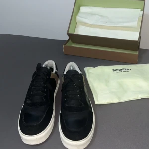 Burberry sneakers med rutigt mönster - Snygga Burberry sneakers i svart skinn och mocka med klassiskt rutigt tyg i beige, svart och vitt på hälen. Vita sulor och svarta snören ger en clean look. Perfekta för dig som vill ha en lyxig streetstyle med ikoniska Burberry-detaljer. Den är strl 42 inga skador bara lite smutsig använd lite men inte mycket tar byten och pris kan diskuteras du får med boxen och allt i bilden