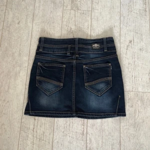 Mörk Lågmidjad jeanskjol från Bigrey - Snygg mörkblå jeanskjol från Bigrey med dubbla knappar framtill och coola fickor både fram och bak. Kjolen har markerade sömmar och bälteshällor, perfekt till en avslappnad streetstyle. Materialet är stretchigt med 98% bomull och 2% lycra. Midjemått 37 cm Längd 35 cm Står M men skulle säga S