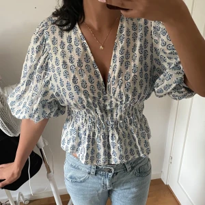Vit och blå blus med puffärm - Supersöt blus i vitt med blått blommigt mönster, djup v-ringning och smockad midja. Puffiga korta ärmar och volangig nederkant ger en romantisk vibe. Perfekt till jeans eller kjol för en fräsch look. Storlek L men passar mig som S i toppar men har större byst 