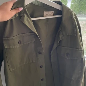 Olivgrön overshirt från Nudie Jeans - Snygg olivgrön overshirt från Nudie Jeans i 100% ekologisk bomull. Skjortan har två stora bröstfickor med knappar och klassisk krage. Perfekt lager-på-lager-plagg med avslappnad passform och stilrena detaljer.