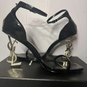 Ysl klackar - Exklusiva svarta sandaler från Saint Laurent i lackat skinn med ikoniska YSL-klackar i guldmetall. Skorna har smal rem över tårna, fyrkantig tå och justerbar ankelrem. Perfekta för dig som vill sticka ut med en lyxig och edgy vibe.