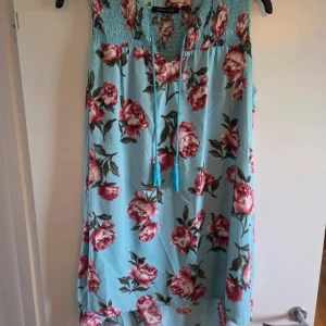 Blommig blå klänning från Mywearwoman - Säljer en ljusblå klänning med rosa och röda blommor från Mywearwoman. Klänningen har smock upptill, snörning vid halsen och är ärmlös. Materialet är mjukt och lätt, perfekt för varma dagar. Passformen är loose och längden är kort.