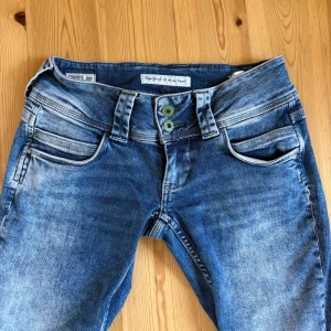Pepe Jeans straight low waist jeans blå - Snygga blå jeans från Pepe Jeans med låg midja och raka ben. Jeansen har slitna detaljer, dubbla gröna knappar i midjan och klassiska bakfickor med broderad våg. Tillverkade i mjukt denim för en bekväm passform. Inga tecken på slitage ! Midjemåttet rakt över är 36cm och innebenslängden är 74cm. 💕💖Säljer jeansen då de är för korta!