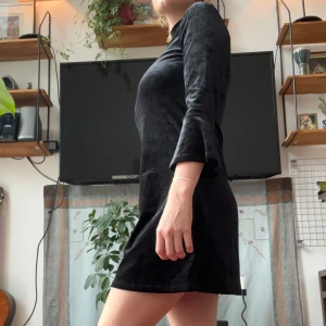 Svart sammet klänning med öppen rygg - Snygg svart kortklänning i mjuk sammet med långärmade trumpetärmar och hög hals. Baksidan har en cool oval öppning som ger extra edge. 