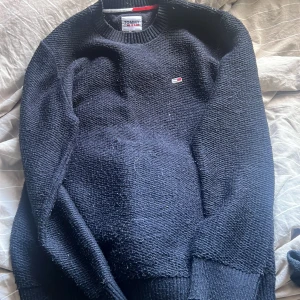 Svart stickad tröja Tommy Jeans - Svart stickad tröja från Tommy Jeans med klassisk rund hals och diskret logga på bröstet. Tröjan har långa ärmar och ribbade muddar vid hals, ärmslut och nederkant. Perfekt för dig som gillar enkel och stilren design med en touch av street.