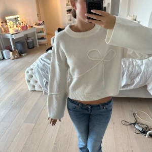 Vit stickad tröja med rosettdetalj H&M - Superfin vit stickad tröja från H&M Divided med en söt rosettdetalj framtill. Tröjan har breda ärmar, ribbade muddar och en croppad passform. Perfekt för lager på lager under höst och vinter. Enkel att styla med jeans eller kjol. Använd 1 gång! 