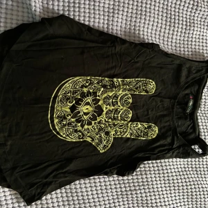 Svart linne med guldigt handtryck - Coolt svart linne med ett stort guldigt tryck av en detaljerad hand med öga på framsidan. Ärmlös modell med rund halsringning, perfekt för en edgy och avslappnad stil.