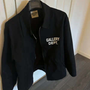 Svart Gallery Dept. overshirt - ALDRIG ANVÄND. Svart overshirt från Gallery Dept.  med vit logga tryckt på bröstet och stor text på ryggen. Jackan har dragkedja framtill, klassisk krage och raka ärmar. Perfekt för dig som gillar streetwear och vill sticka ut med en clean look. STORLEK S 