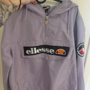 Ljuslila vindjacka från Ellesse - Snygg ljuslila vindjacka från Ellesse med stor logga framtill och patch på ärmen. Jackan har huva med dragsko, halv dragkedja och praktiska fickor. Använd fåtal gånger