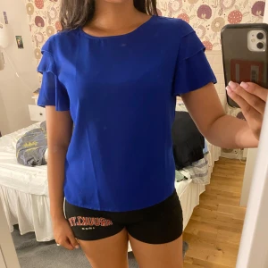 Kornblå blus med volangärm från SHEIN - Säljer en kornblå blus från SHEIN i storlek S. Blusen har korta ärmar med dubbla volangdetaljer och rund halsringning. Materialet är lätt och flowy, perfekt för en fräsch look. Enkel att matcha med både jeans och kjol.