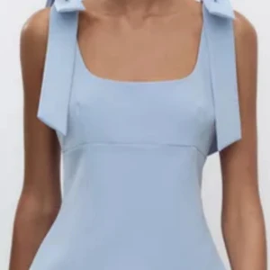 Ljusblå satinlinnetopp från Zara - Supersöt ljusblå linne-topp från Zara i satin med breda axelband som knyts i stora rosetter. Toppen har en figurnära passform och fyrkantig ringning som ger en modern och trendig look. Perfekt att styla med jeans eller kjol för en fräsch vibe.