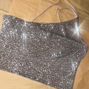 Glittrig silver one shoulder topp - Säljer en glittrig silverfärgad topp med paljetter och smal axelrem. Toppen har en snygg one shoulder-design och är croppad, perfekt för att sticka ut. Materialet är syntet och insidan är mjukare tyg. Superfin till fest eller när du vill glänsa extra. Använd en gång 