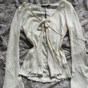 Vit omlottopp med knyt från Bik Bok - Supersöt vit omlottopp från Bik Bok i storlek S. Toppen har långa ärmar, v-ringning och knyt framtill. Materialet är strukturerat och ger en trendig känsla. Perfekt att styla med jeans eller kjol för en fräsch look.